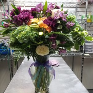 Just Because Bouquet - A vibrant floral arrangement in a glass vase features purple, orange, and green flowers along with lush greenery. The vase is adorned with a purple ribbon.