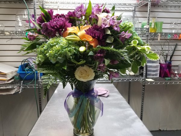 Just Because Bouquet - A vibrant floral arrangement in a glass vase features purple, orange, and green flowers along with lush greenery. The vase is adorned with a purple ribbon.