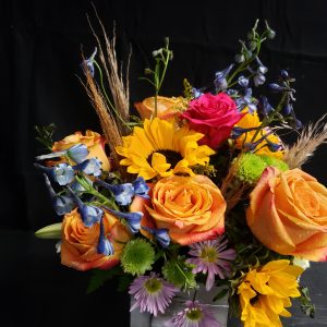 A vibrant floral arrangement featuring orange roses, pink rose, yellow sunflowers, blue delphiniums, and purple daisies, set against a dark background. The flowers are arranged in a rectangular, metallic container.