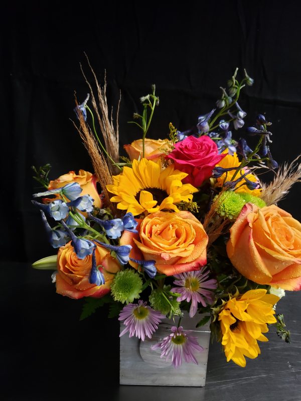 A vibrant floral arrangement featuring orange roses, pink rose, yellow sunflowers, blue delphiniums, and purple daisies, set against a dark background. The flowers are arranged in a rectangular, metallic container.