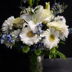 A vibrant bouquet of white lilies, daisies, and small blue flowers in a clear glass vase, set against a dark background.