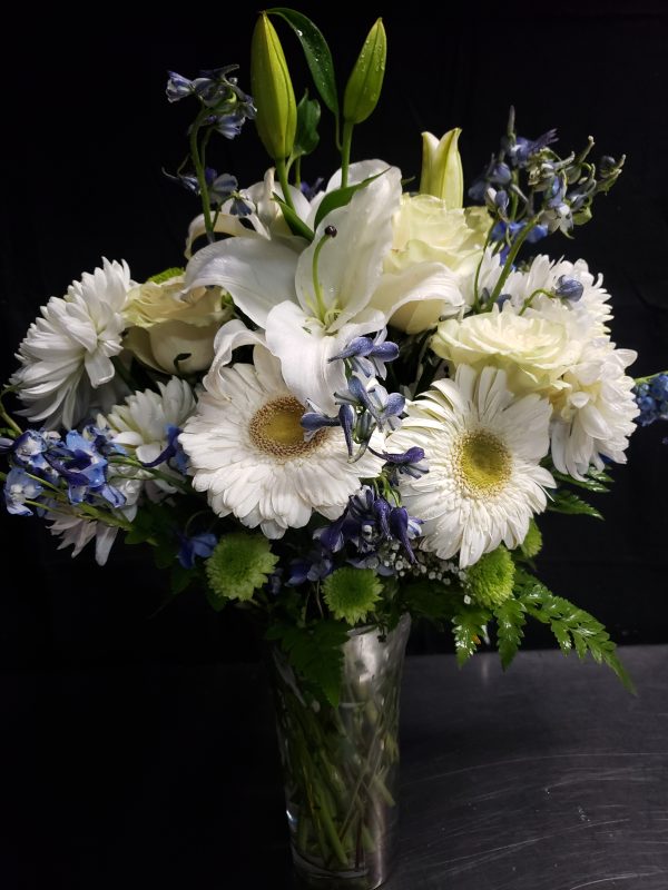 A vibrant bouquet of white lilies, daisies, and small blue flowers in a clear glass vase, set against a dark background.