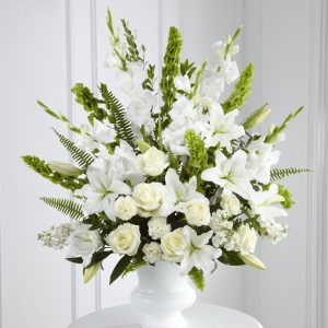 A white flower arrangement in a pedestal vase, featuring roses, lilies, and gladiolus, accented with green ferns and leaves, set against a light background.