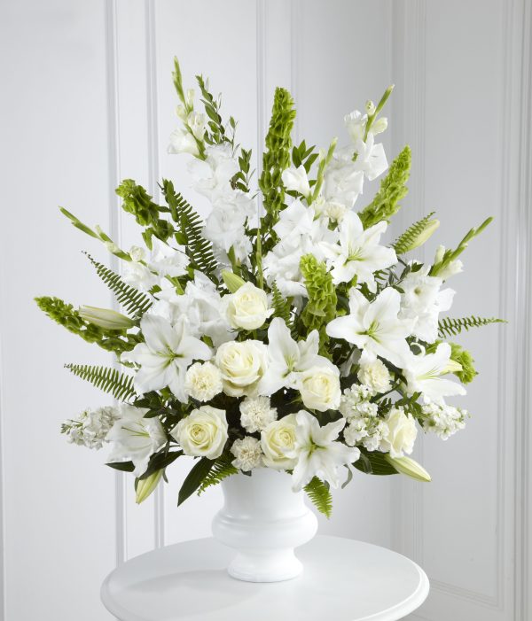 A white flower arrangement in a pedestal vase, featuring roses, lilies, and gladiolus, accented with green ferns and leaves, set against a light background.