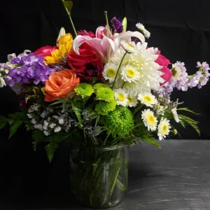 A vibrant floral arrangement in a clear glass vase against a dark background. The bouquet includes white daisies, pink lilies, orange and pink roses, and green foliage, creating a colorful and lively display.