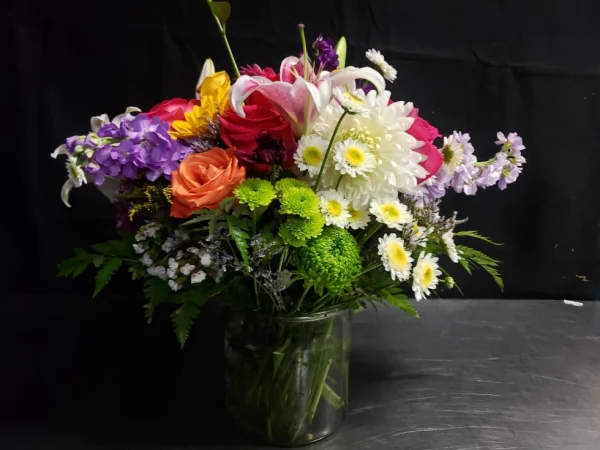 A vibrant floral arrangement in a clear glass vase against a dark background. The bouquet includes white daisies, pink lilies, orange and pink roses, and green foliage, creating a colorful and lively display.