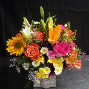 A vibrant floral arrangement featuring orange roses, sunflowers, lilies, pink roses, and various wildflowers with lush greenery in a rectangular silver vase. The blossoms are brightly colored, creating a cheerful display against a dark background.