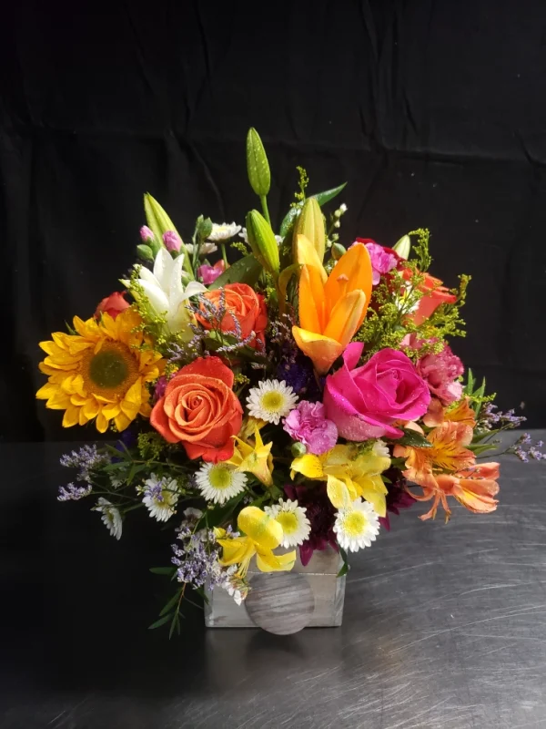 A vibrant floral arrangement featuring orange roses, sunflowers, lilies, pink roses, and various wildflowers with lush greenery in a rectangular silver vase. The blossoms are brightly colored, creating a cheerful display against a dark background.