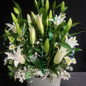 A bouquet of white lilies and daisies with green leaves and decorative ribbons, arranged in a light gray pot against a dark background.