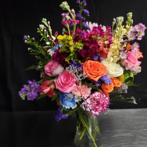 A vibrant bouquet featuring a variety of blooms, including roses, carnations, and wildflowers in pink, orange, purple, and blue hues, arranged in a clear glass vase against a dark background.