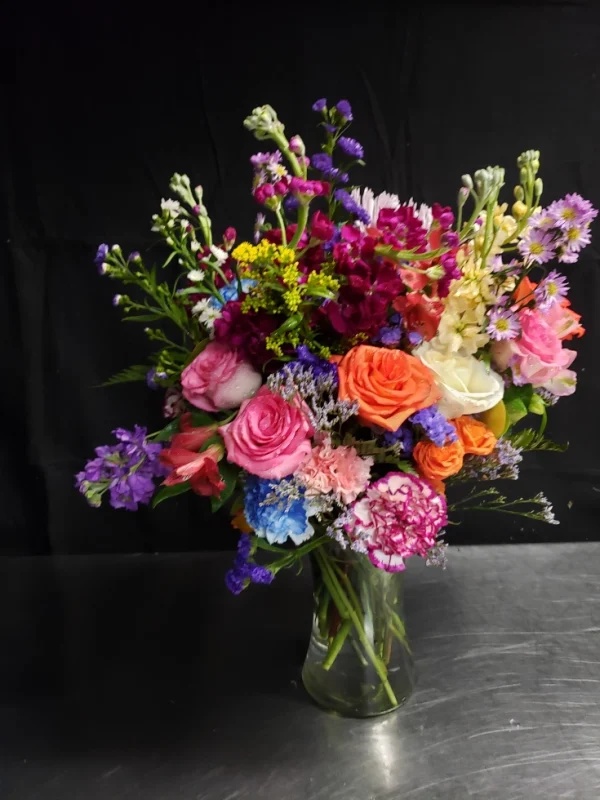 A vibrant bouquet featuring a variety of blooms, including roses, carnations, and wildflowers in pink, orange, purple, and blue hues, arranged in a clear glass vase against a dark background.