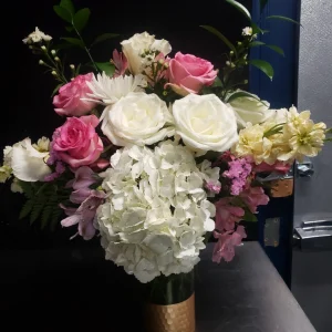 A bouquet in a gold vase featuring white hydrangeas, pink roses, white roses, and light pink and white flowers, accented with greenery, set against a dark background.