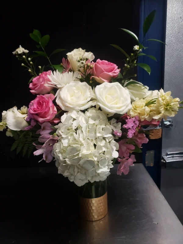 A bouquet in a gold vase featuring white hydrangeas, pink roses, white roses, and light pink and white flowers, accented with greenery, set against a dark background.