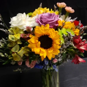 A vibrant bouquet in a glass vase featuring sunflowers, roses, and assorted colorful blooms against a dark background.