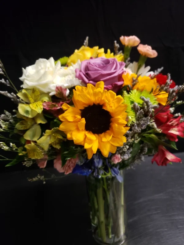 A vibrant bouquet in a glass vase featuring sunflowers, roses, and assorted colorful blooms against a dark background.