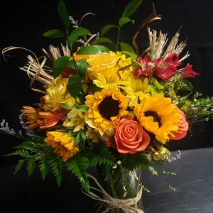 A vibrant bouquet featuring sunflowers, orange roses, and yellow daisies, accented with wheat stalks and green foliage, arranged in a glass vase against a dark background.