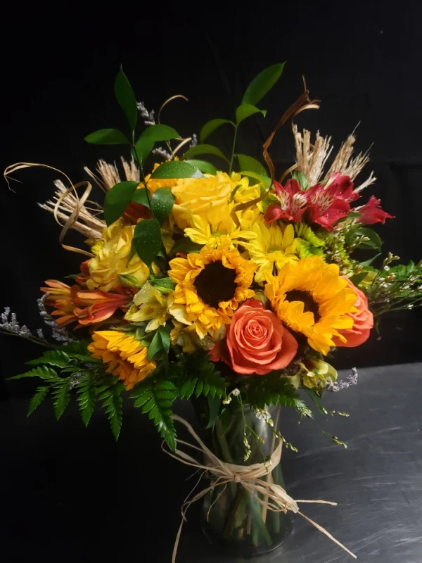 A vibrant bouquet featuring sunflowers, orange roses, and yellow daisies, accented with wheat stalks and green foliage, arranged in a glass vase against a dark background.