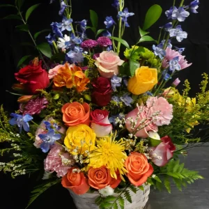 A vibrant floral arrangement featuring roses, chrysanthemums, and delphiniums in shades of red, pink, yellow, and blue, accented with lush greenery, set in a white basket against a dark background.