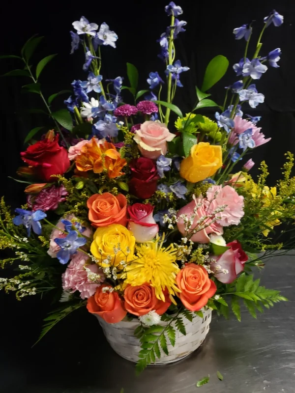 A vibrant floral arrangement featuring roses, chrysanthemums, and delphiniums in shades of red, pink, yellow, and blue, accented with lush greenery, set in a white basket against a dark background.