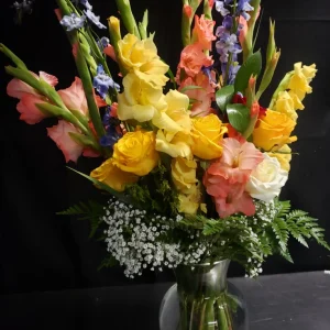 A vibrant floral arrangement in a glass vase, featuring yellow roses, pink and orange gladiolus, and sprigs of baby's breath, set against a dark background.