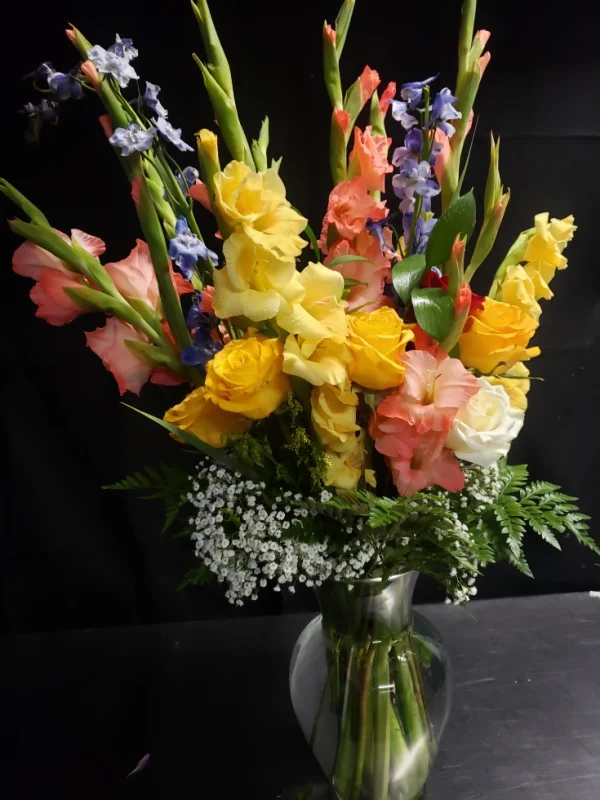 A vibrant floral arrangement in a glass vase, featuring yellow roses, pink and orange gladiolus, and sprigs of baby's breath, set against a dark background.