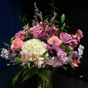 A vibrant bouquet of pink and purple roses, white hydrangeas, and assorted greenery arranged in a clear glass vase against a dark background.