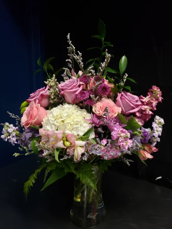 A vibrant bouquet of pink and purple roses, white hydrangeas, and assorted greenery arranged in a clear glass vase against a dark background.