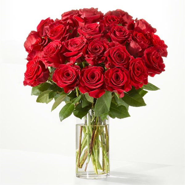 A bouquet of vibrant red roses arranged in a clear glass vase against a plain white background. The roses are in full bloom, with lush green leaves adding contrast to the deep red petals.